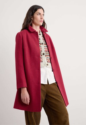 Red wool coat with a high collar, textured fabric, and button closure, worn over a multi-patterned sweater and white shirt, paired with brown cords.