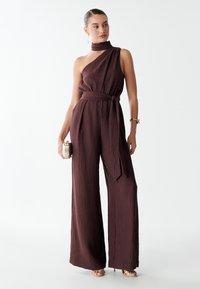 Combinaison une épaule bordeaux en tissu texturé, avec une coupe large, une taille nouée et un petit foulard au cou, assortie à une pochette métallique.
