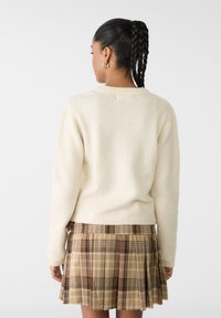 Cremefarvet, fuzzy sweater med lange ærmer, ledsaget af en brun plaid plisseret nederdel i beige og mørkere nuancer.