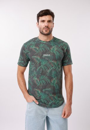 Homme portant un t-shirt à imprimé feuilles tropicales vert foncé avec le texte "IBIZA", jean bleu clair, debout devant un fond blanc uni.