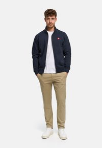 Marinblå huvtröja med dragkedja och en liten röd logotyp, kombinerad med beige chinos och vita sneakers. Modellen står med händerna i fickorna.