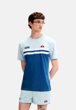 Joven con gafas que lleva una camiseta azul y gris claro de ellesse y pantalones cortos a juego, de pie con las manos detrás de la espalda.