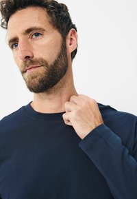 Marineblaue Langarmshirt aus weichem Baumwollmaterial, Rundhalsausschnitt und taillierte Ärmel, mit sichtbaren Nähdetails. Das Model passt sanft den Kragen an.