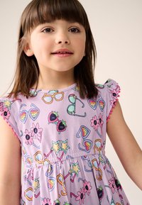 Fialové šaty s barevnými potisky slunečních brýlí, květin a jahod, se sukní do zvonu a lemováním střapci na rukávech.