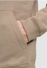 Beżowa bluza z kapturem z kieszenią typu kangur i ściągaczami. Miękka, gładka faktura materiału z widocznymi detalami szwów oraz małą metką z logo marki.