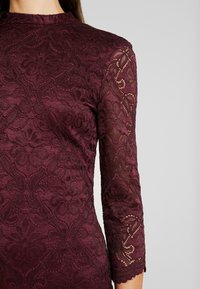 Femme portant une robe en dentelle bordeaux ajustée à manches trois-quarts et motif floral, montrée du cou aux hanches sur un fond uni.