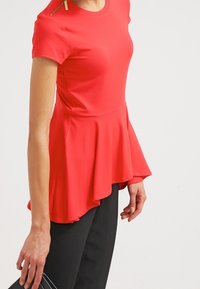 Röd peplumtopp med korta ärmar, som har en hög halsringning och en dragkedjedetalj på axeln. Slät tyg, utställd nederkant, kombinerad med svarta byxor.