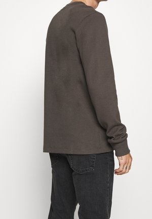 Ein Mann trägt ein langärmeliges dunkelbraunes Hemd und schwarze Jeans, steht leicht vom Kamerawinkel weggedreht vor einem weißen Hintergrund.