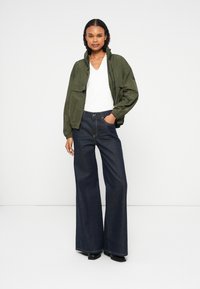 Πράσινη bomber jacket με ψηλό γιακά, φορεμένη πάνω από ένα λευκό top με V-neck. Μπλε παντελόνια σε φαρδύ στιλ και μαύρες πλατφόρμες. Καθαρό φόντο.