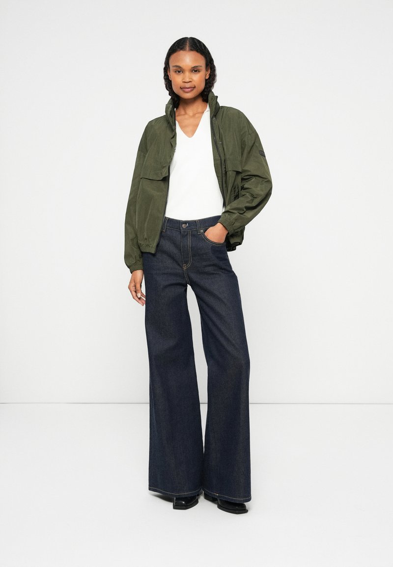 Grüne Bomberjacke mit Stehkragen, getragen über einem weißen V-Ausschnitt-Oberteil. Hellblaue, weit geschnittene Jeans und schwarze Plateauschuhe. Sauberer Hintergrund.