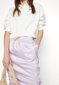 Sudadera blanca de gran tamaño con cuello y logo negro de Nike, combinada con falda ligera púrpura brillante, fruncida en los lados con logo.