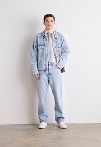 Lichtblauwe denimjas en -broek, gecombineerd met een beige shirt en stropdas, en witte sneakers. Model staat tegen een effen achtergrond.