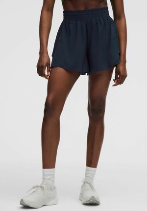 Person iført mørkeblå træningsshorts, en grøn crop top, hvide sokker og hvide sneakers, stående mod en ensfarvet baggrund.