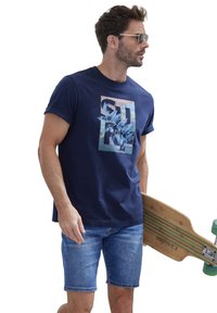 Dunkelblaues Baumwoll-T-Shirt mit einem bunten floralen Grafikdesign, kombiniert mit hellen Jeansshorts. Das Model hält ein Holzskateboard.