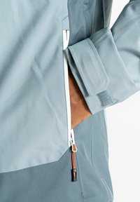 Veste bleu clair avec une poche zippée et des accessoires oranges et noirs. Texture lisse, coupe ajustée avec des poignets réglables.