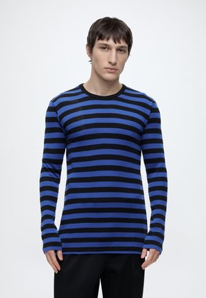 Ung mand iført en blå og sort langærmet t-shirt med vandrette striber, stående mod en ensartet hvid baggrund.