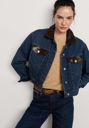 Mujer con chaqueta de mezclilla azul con cuello y bolsillos de cuero marrón oscuro, suéter beige y vaqueros azules con cinturón marrón.