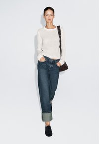 Sweter w kolorze kremowym z dzianiny, luźne niebieskie dżinsy z podwiniętymi mankietami, ciemna torba, czarne buty; model stoi na jednolitym jasnym tle.