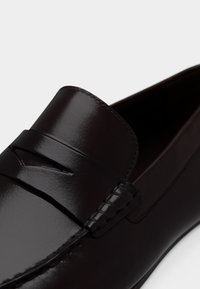 Gros plan sur un mocassin en cuir brun foncé poli, avec un bout mocassin et une bride traversant le cou-de-pied.