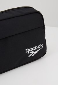 Pochette en tissu noir avec une fermeture éclair et le logo Reebok blanc brodé sur la poche avant, posée sur une surface claire.