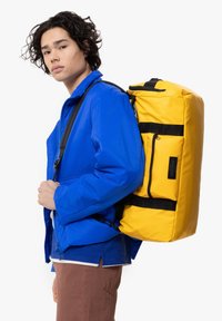 Mochila amarilla con acentos negros, fabricada en material duradero, forma rectangular, correas ajustables y un bolsillo exterior con cremallera.