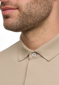 Polo beige con tessuto testurizzato, dotato di un colletto a punta e di un bottone bianco, catturato da una prospettiva ravvicinata.