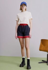 Camiseta corta de manga en color crema, pantalones cortos negros y rojos con detalles en blanco, gorra azul y botas negras con cordones y suelas de plataforma.