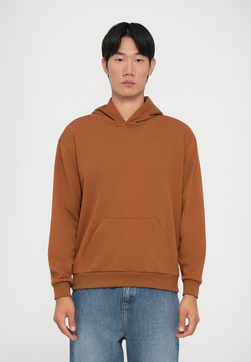 Sweatshirt à capuche marron en tissu doux, doté d'une poche avant et de poignets côtelés. Porté avec un jean bleu clair déchiré.