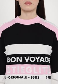 Femme portant un t-shirt rayé noir, blanc et rose avec les inscriptions "BON VOYAGE", "STIEGLITZ" et "ORIGINALE - 1988" imprimées sur le devant.