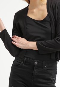 Svart cardigan i mjukt material med cropped design och långa ärmar, bärs över en enkel svart linne, parat med slitna svarta jeans.