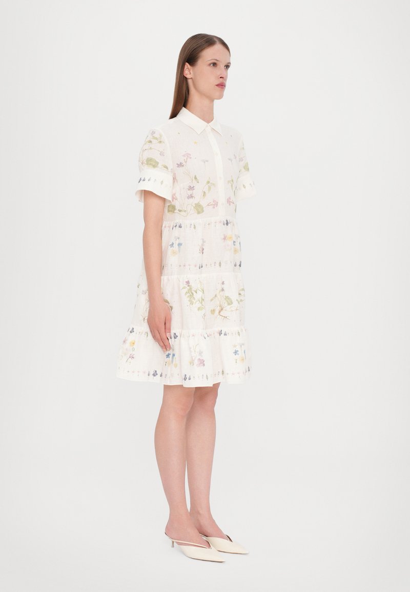 Robe blanche à manches courtes avec broderies florales, col rabattu, design en couches et ourlet avec volants. Portée avec des chaussures beige à bout pointu.
