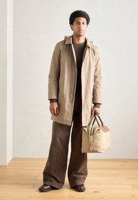 Bézs esőkabát kapucnival, barna texturált pulóver, széles barna nadrág és világosbarna duffel táska sötét részletekkel.