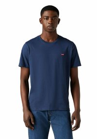 Giovane uomo che indossa una maglietta Levi's blu navy semplice e jeans blu, in piedi contro uno sfondo bianco con un'espressione neutra.