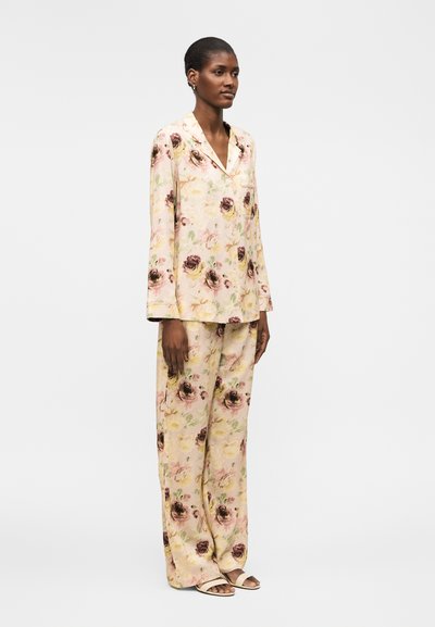 Naine kannab lahtist pika varrukaga lillemustriga pidžaamaülaosa ja sobivaid laiapõhjalisi pükse avatud varbaga sandaalidega, seisab ühevärvilise tausta ees.