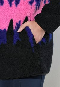 Fleece mikina má růžovo-fialový tie-dye vzor na černém podkladu, s boční kapsou a strukturovanou tkaninou.