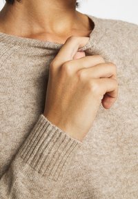 Personne tirant le col d'un pull beige en tricot près du cou nu avec la main droite, montrant la texture du tissu et le poignet côtelé.