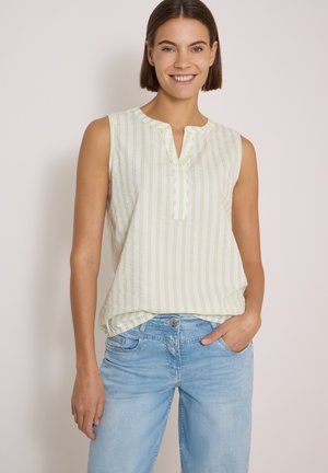 Donna sorridente con capelli castani corti che indossa un top bianco senza maniche a righe verdi e jeans azzurri, mano in tasca.
