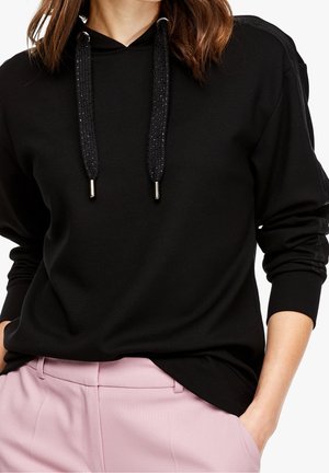 Hoodie noire avec des cordons de serrage noirs brillants, présentant une coupe décontractée et des manches longues. Associé à un pantalon rose clair pour le contraste.