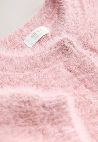 Pulover roz pufos cu textură moale, având un decolteu rotund și o etichetă pe care scrie "NEXT, MADE IN CHINA".