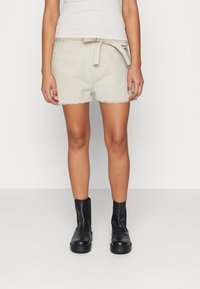 Shorts de mezclilla beige con dobladillo deshilachado, cinturón en la cintura y bolsillos laterales, combinados con botas negras hasta el tobillo. Textura suave y diseño casual.