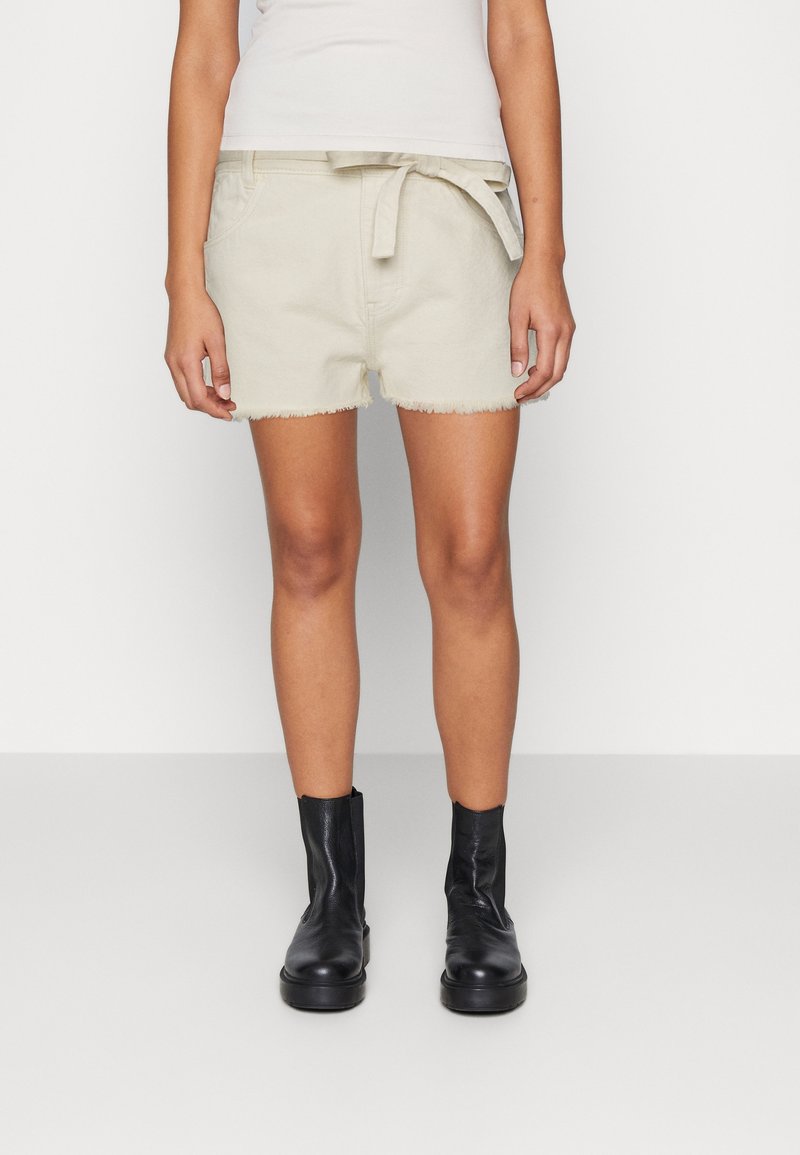 Shorts de mezclilla beige con dobladillo deshilachado, cinturón en la cintura y bolsillos laterales, combinados con botas negras hasta el tobillo. Textura suave y diseño casual.