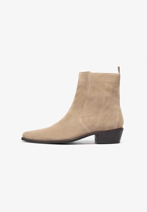 Beige suède enkellaars met een puntige neus, lage blokhak en trekkoord aan de achterkant, weergegeven in zijprofiel.