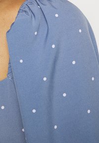 Vaaleansininen kangas, jossa on valkoisia pilkkuja, olkapäissä rypytetty ommel ja sileä tekstuuri.