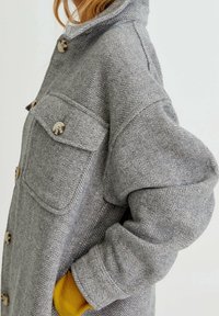 Femme portant un manteau gris à motif chevrons avec des boutons bruns et une manche jaune visible au poignet, debout devant un fond uni.