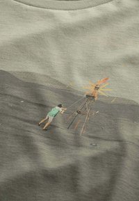 Light green cotton t-shirt featuring a mural of a person operating a wind turbine against a dark gray background.