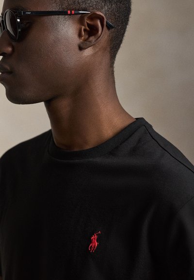 Άνδρας που φοράει μαύρα γυαλιά ηλίου και μαύρο πουκάμισο με μικρό κόκκινο κεντητό λογότυπο παίκτη πόλο στο στήθος.