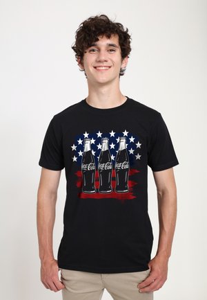 Black short-sleeve t-shirt featuring three vintage Coca-Cola bottles on a red and blue star background. Fabric appears smooth, design colorful.