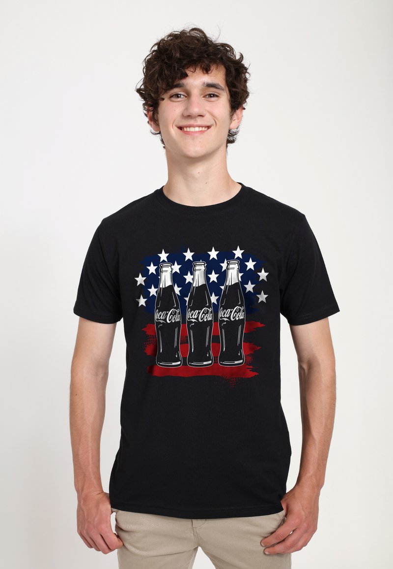 Schwarzes T-Shirt mit kurzen Ärmeln, das drei Vintage-Coca-Cola-Flaschen vor einem rot-blauen Sternenhintergrund zeigt. Der Stoff wirkt glatt, das Design farbenfroh.
