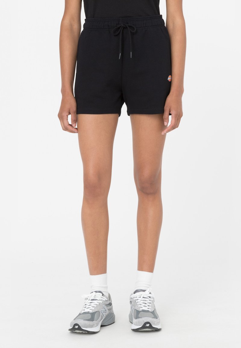 Pantalones cortos de algodón negros con banda elástica en la cintura y cordón. Presentan un pequeño logo en el muslo. Combinados con zapatillas de running grises y calcetines blancos.