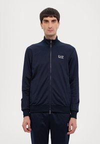 Jachetă bleumarin cu fermoar, realizată din țesătură netedă, cu guler înalt, manșete ribbate și logo subtil EA7. Dispune de un design elegant și ajustat.